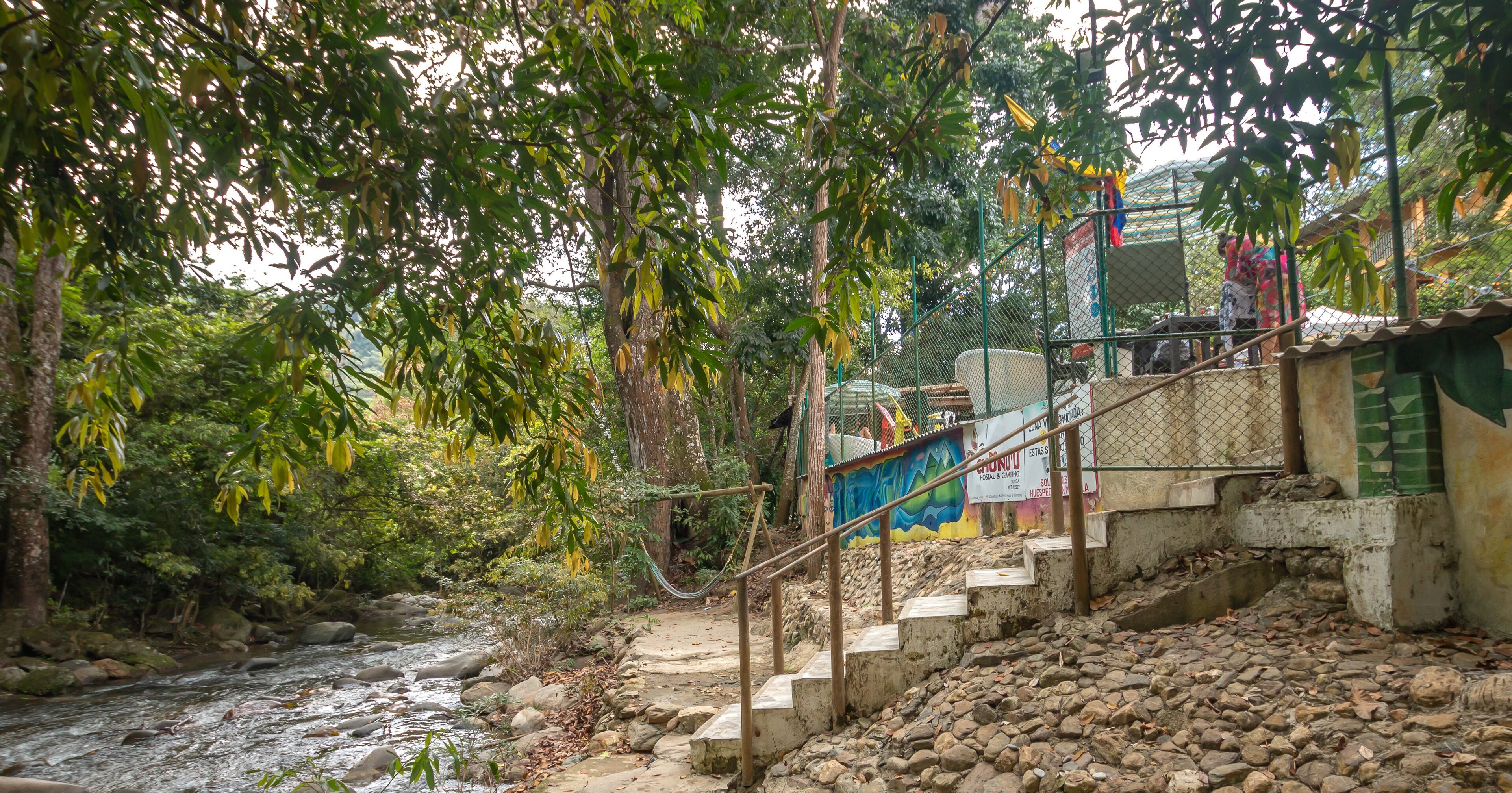 Una escalera de piedra desciende hacia un río, rodeada de abundante vegetación y un mural en una cerca.