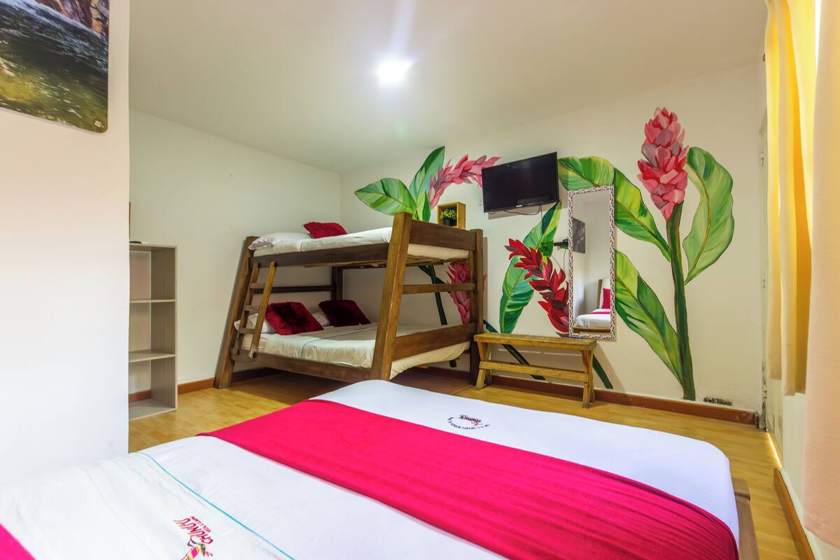 Una habitación con una cama individual superpuesta, una cama plana con una manta roja, un televisor montado en la pared y un espejo con diseño floral.
