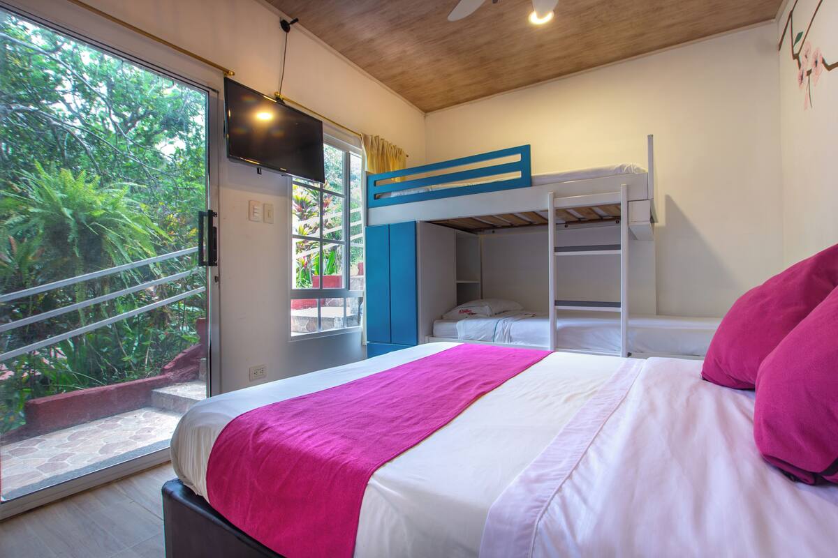 Un dormitorio con una cama litera, un ventanal con vistas a zonas verdes y una cama con una colcha rosa.