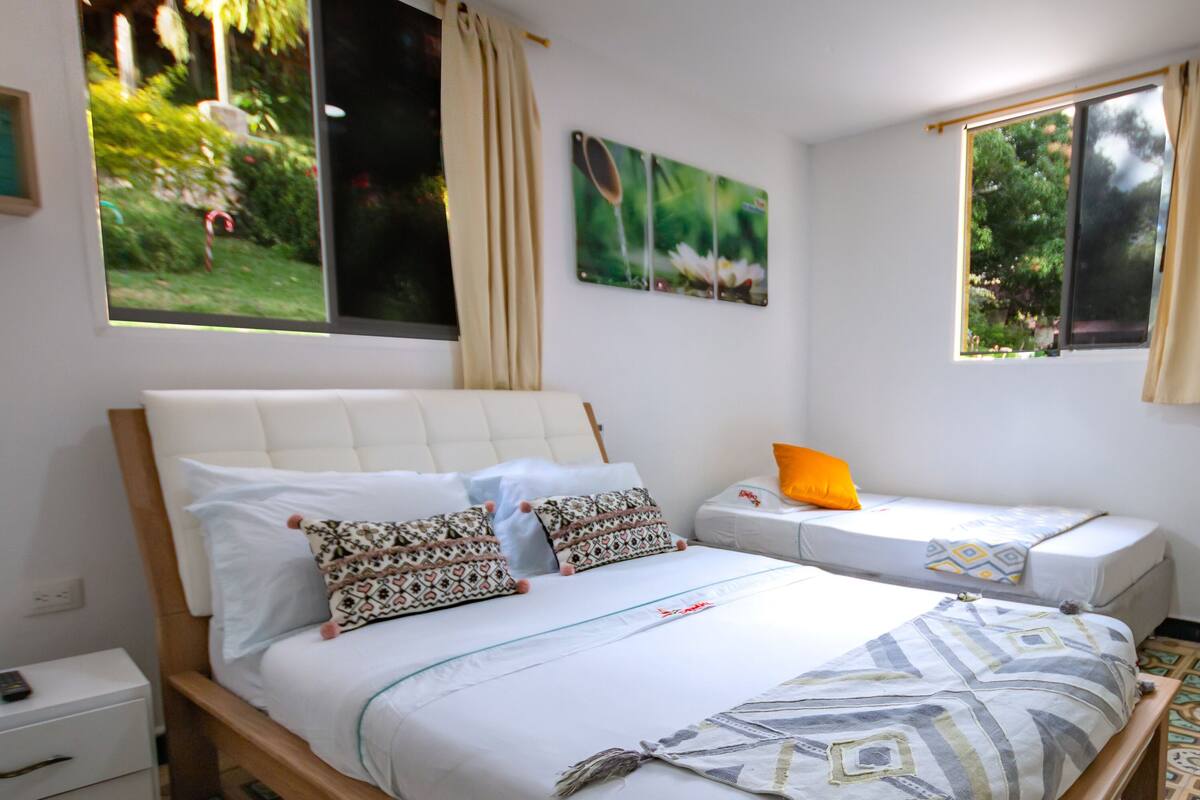 Un dormitorio con una cama grande, una cama más pequeña, una mesita de noche y un taburete de madera. Hay dos ventanas con cortinas, un televisor montado en la pared y una obra de arte con temática de bicicletas.