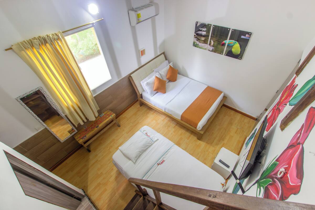 Una habitación con cama, ventana con cortinas, una mesita y televisor montado en la pared.