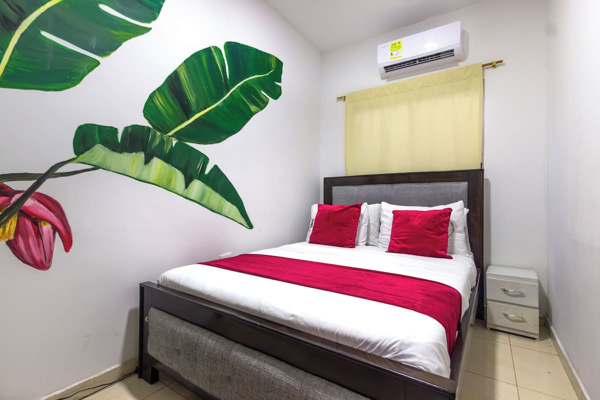 Un dormitorio con cama, mesita de noche, aire acondicionado instalado en la pared y un mural de plantas.