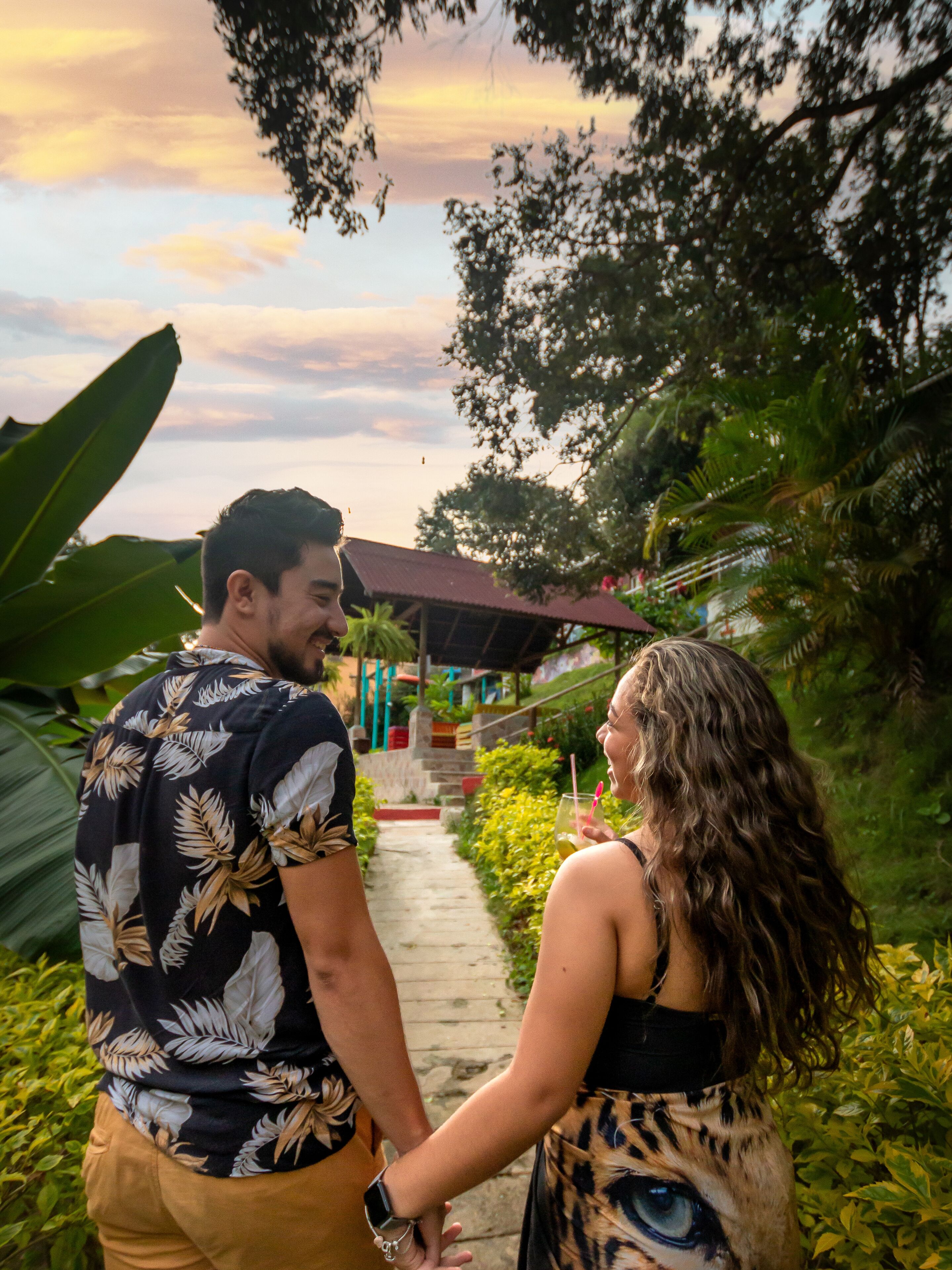 A couple holding hands on a pathway surrounded by lush greenery and a building with a red roof.