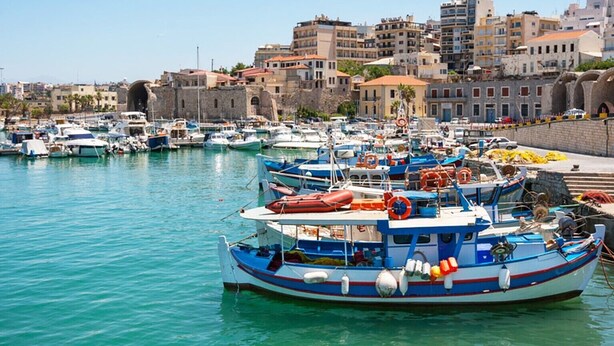 A marina with multiple boats docked, a stone building in the background, and a clear blue sky. - Orange Central Studio / Heraklion Crete  (Ηράκλειο)