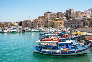 A marina with multiple boats docked, a stone building in the background, and a clear blue sky.