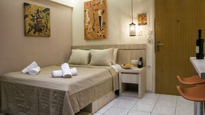 A hotel room with a bed, pillows, a nightstand, a bottle of water, a bowl of fruit, and two paintings on the wall. - Orange Central Studio / Heraklion Crete  (Ηράκλειο)