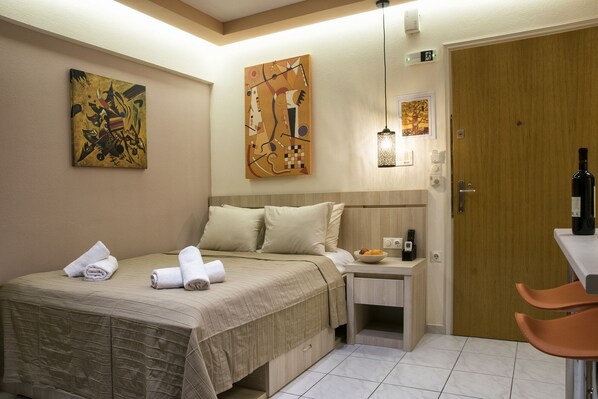 A hotel room with a bed, pillows, a nightstand, a bottle of water, a bowl of fruit, and two paintings on the wall.