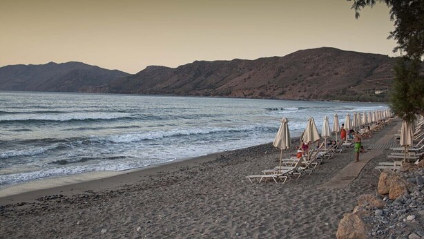 Una playa con hileras de tumbonas y sombrillas, terreno rocoso y montañas al fondo.