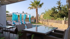 Un patio con una mesa de vidrio y sillas de mimbre, una piscina con cortinas azules y un muro de piedra con una palmera.