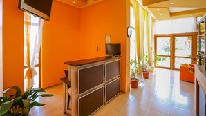 Ett ljust rum med orange väggar, en tv på ett tv-bord, ett skrivbord och ett stort fönster med gardiner.