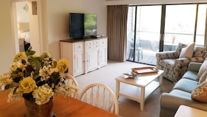 Ein Wohnzimmer mit Fernseher, einer Couch, einem Esstisch mit Blumen und einem Balkon mit Blick.