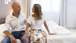 A couple sitting on a bed in a hotel room with a neatly made bed and a stack of towels. - Anda e Rianda (Campi Bisenzio)