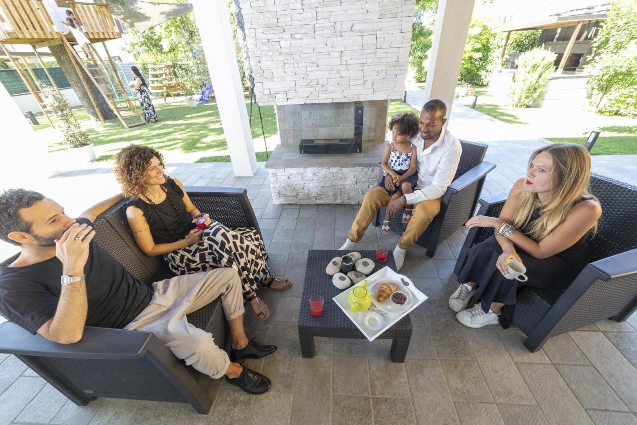 Un grupo de personas sentadas alrededor de una mesa con una bandeja de comida y bebidas en un patio con chimenea de piedra.