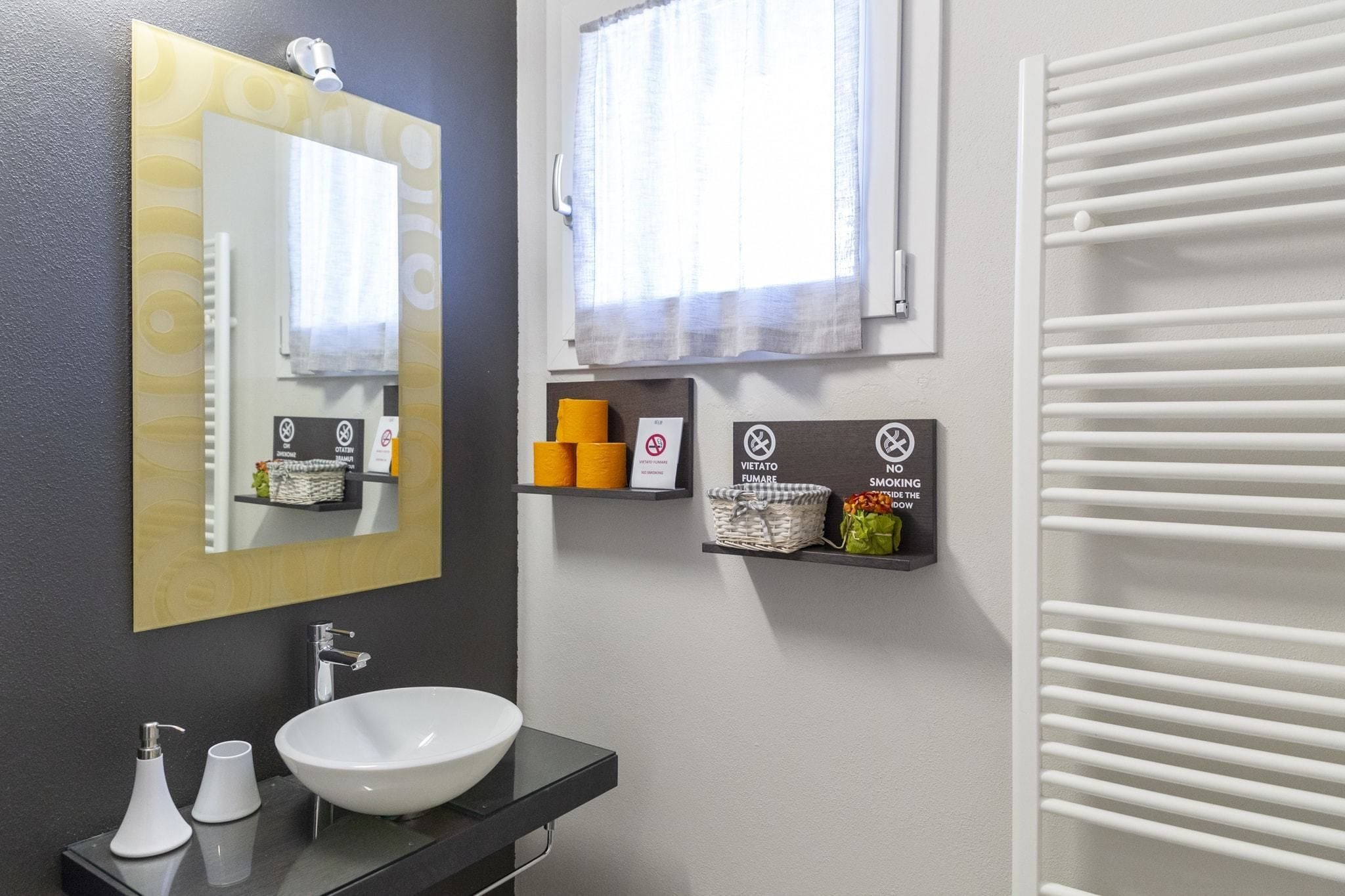 A modern bathroom with a large mirror, a white sink, and a towel rack. There are two shelves with toiletries and a sign indicating no smoking.