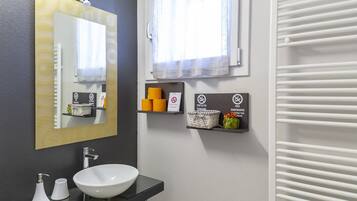 A modern bathroom with a large mirror, a white sink, and a towel rack. There are two shelves with toiletries and a sign indicating no smoking.