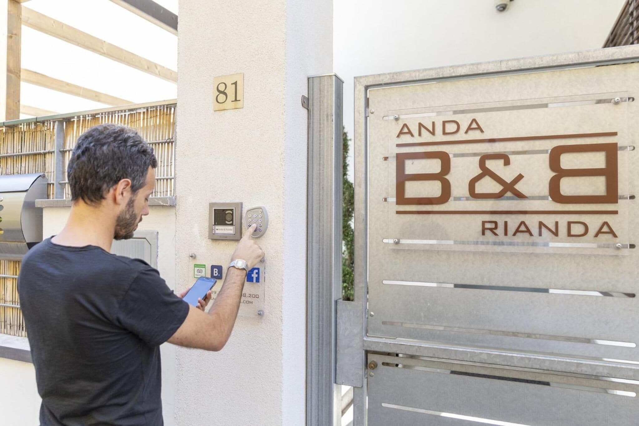 A man using a keypad and a mobile device at a hotel entrance with a metal gate and a sign that reads "ANDA B&B RIANDA".