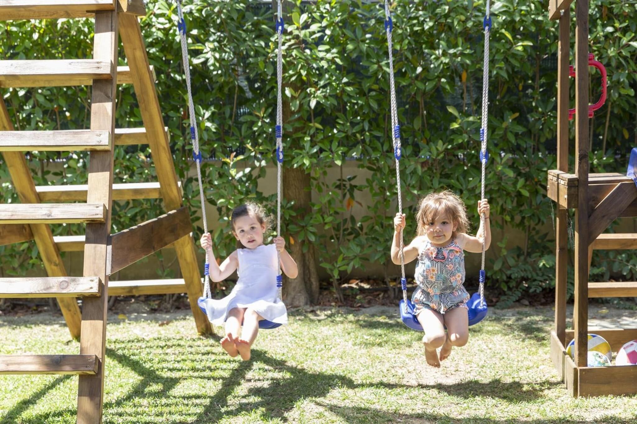 Two children on swings in a wooden play area with a ladder and a slide.