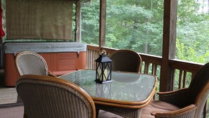 A covered outdoor seating area with wicker chairs and a glass table, overlooking a forested area.