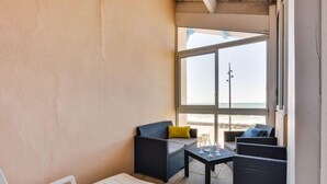 Ett rum med ett stort fönster med utsikt över stranden, inrett med vita loungestolar, en svart soffa med gula kuddar och ett litet bord med vinglas.
