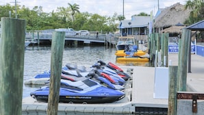 A jet ski rental area with multiple jet skis docked at a pier, a small boat, and a thatched-roof building in the background.