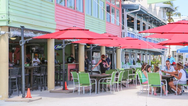 An outdoor dining area with green chairs and red umbrellas, a bar area, and a colorful building with multiple windows.