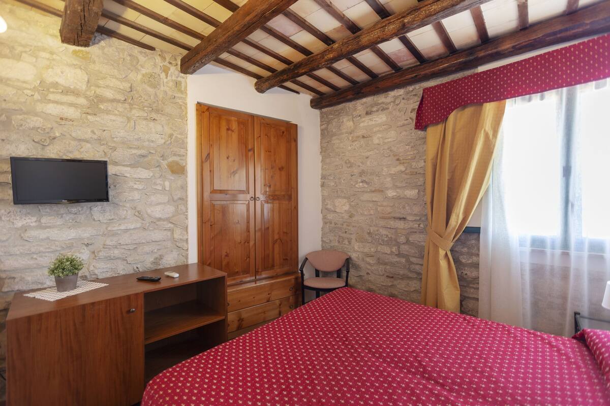A bedroom with a stone wall, wooden wardrobe, a bed with a red bedspread, a television mounted on the wall, and a window with curtains.