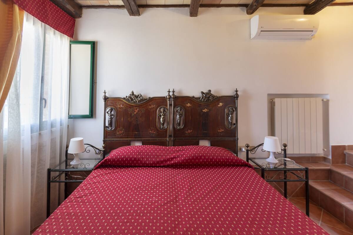 A bedroom with a wooden bed, two bedside tables with lamps, a window with curtains, and a radiator.