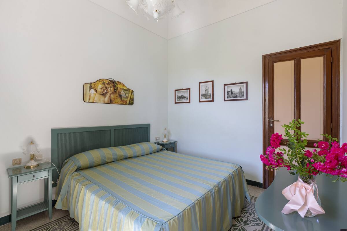 A bedroom with a bed, bedside tables, a nightstand, a wall-mounted painting, framed pictures, and a vase with flowers.