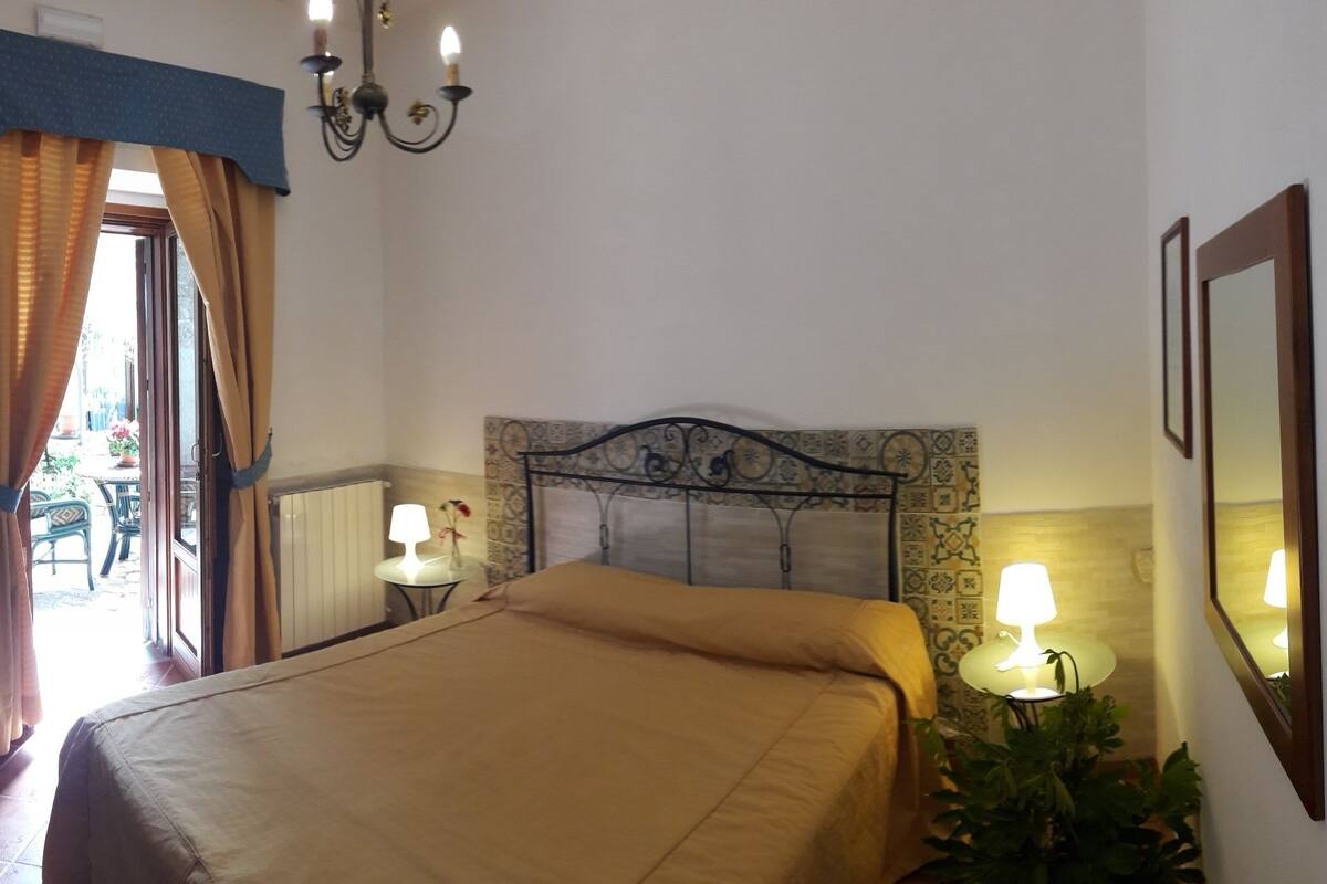 A bedroom with a bed, two bedside lamps, a chandelier, a window with curtains, a radiator, and a wall with a framed picture.