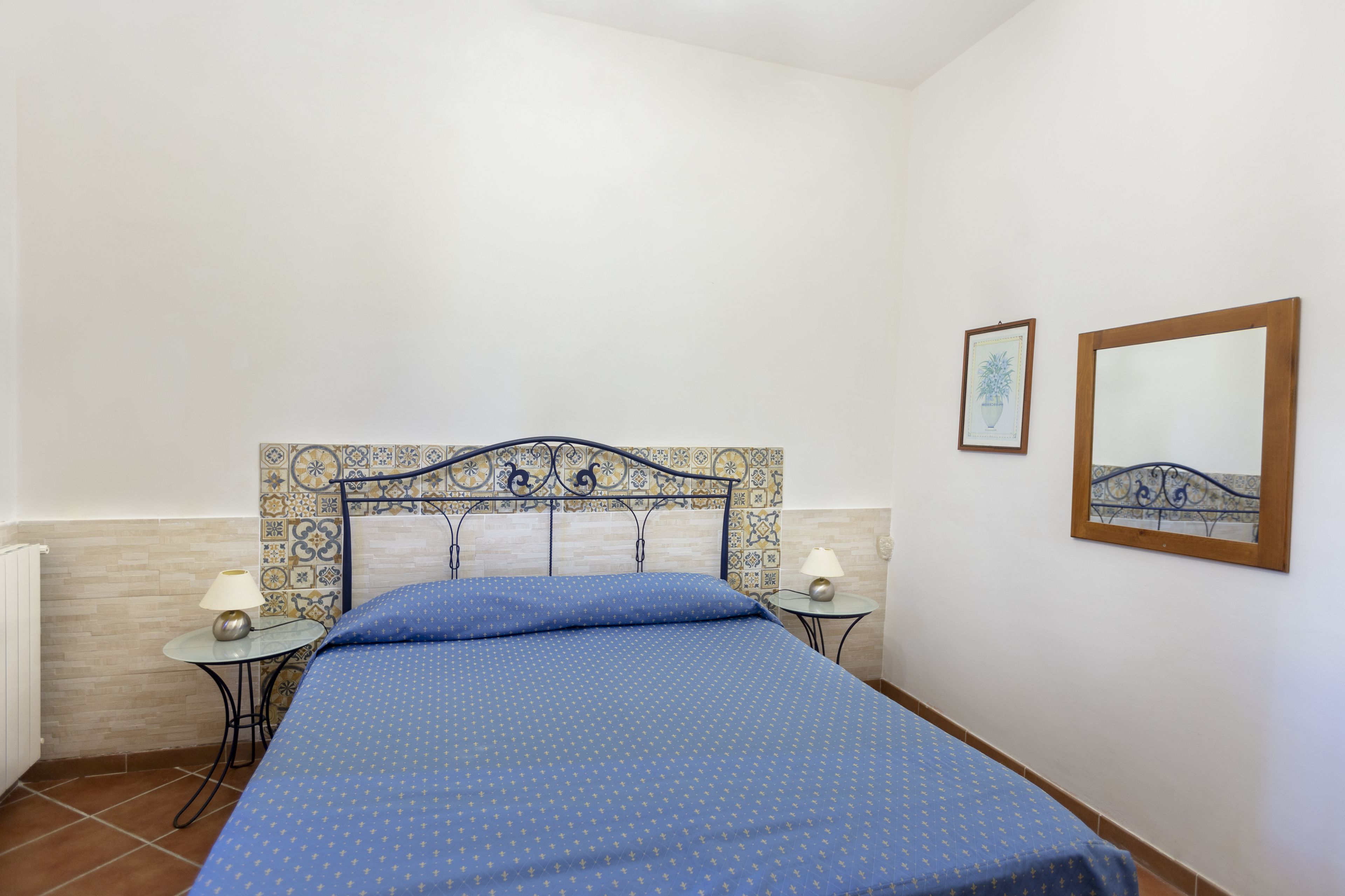 A bedroom with a bed, two bedside tables, a wall with a decorative tile pattern, and two framed pictures.