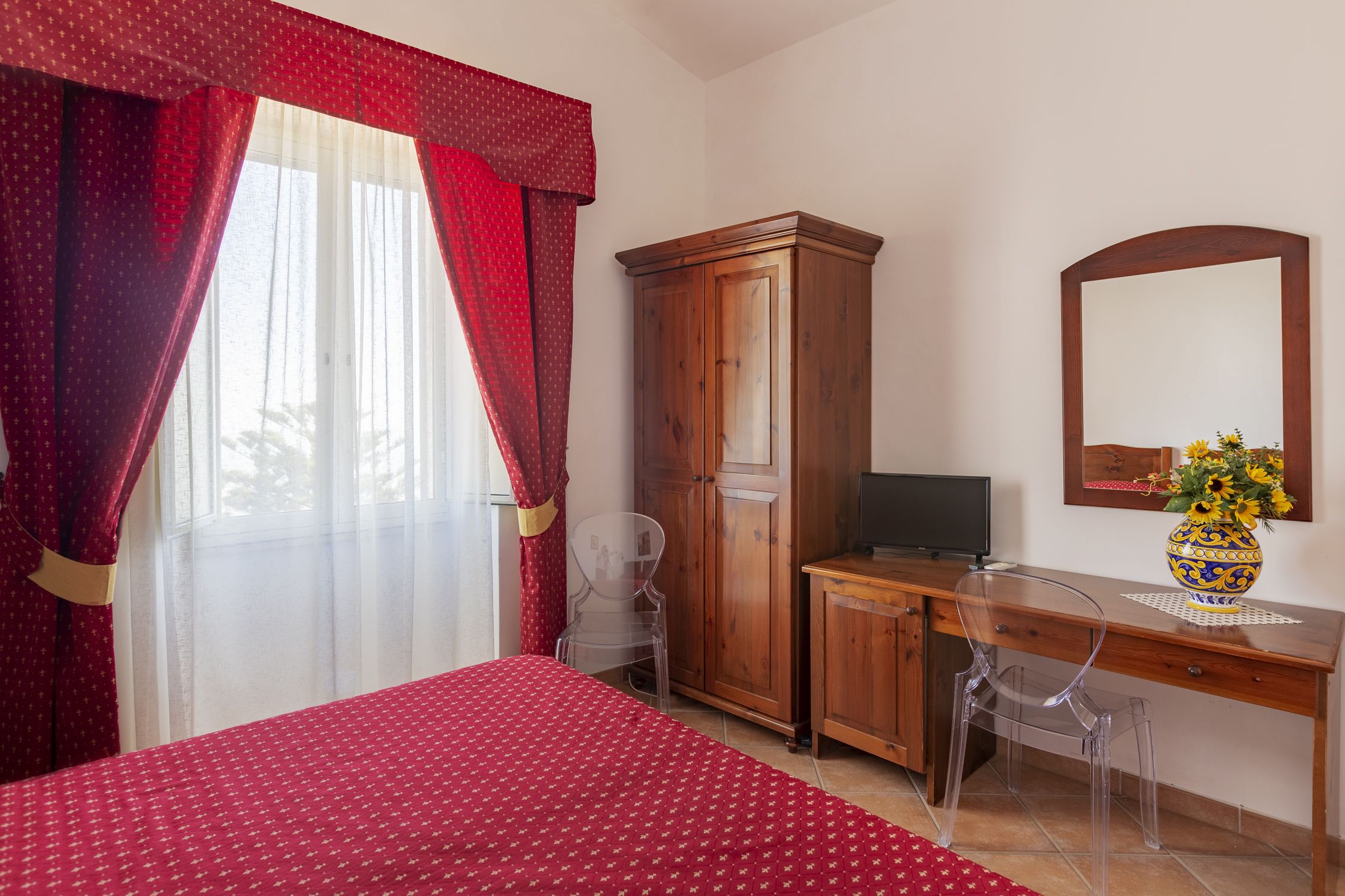 Un dormitorio con cama, un escritorio de madera, una silla, un armario, televisor y un jarrón con flores.