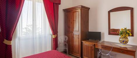 Un dormitorio con cama, un escritorio de madera, una silla, un armario, televisor y un jarrón con flores.