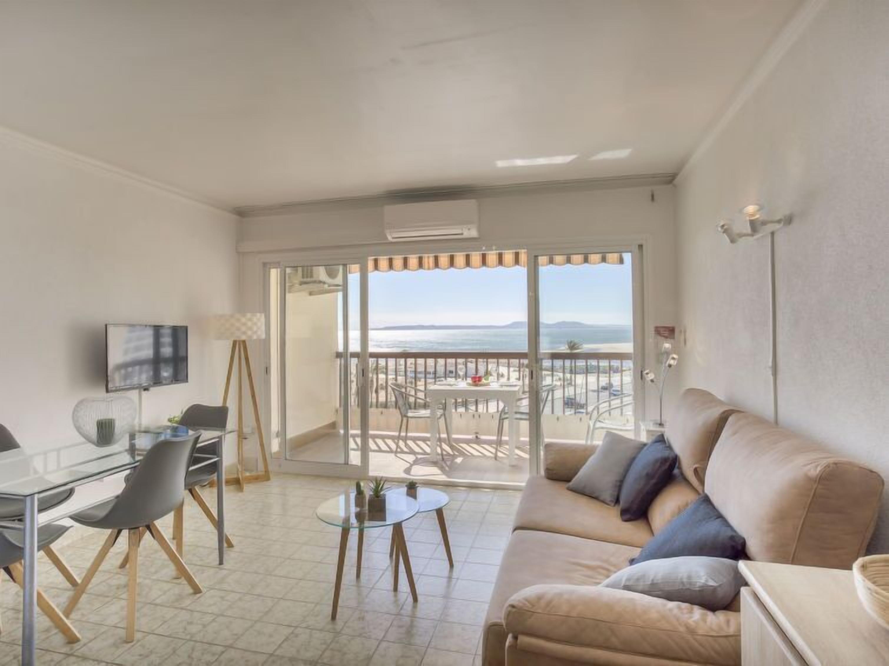 Una sala de estar con un sofá, una mesita de vidrio y un televisor. Hay un balcón con vista al mar.