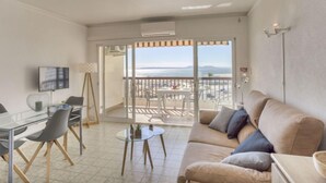 Un salon avec un canapé, une table en verre et un téléviseur. Il y a un balcon avec vue sur la mer.