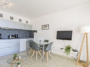 A modern kitchen with grey cabinets, a built-in oven, and a coffee machine. There is a dining area with a glass table and chairs, and a flat-screen TV mounted on the wall.