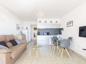 A modern living room with a sofa and a glass coffee table, a kitchen with white cabinets, and a dining area with a table and chairs.