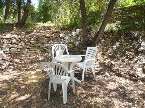 A stone wall, two white plastic chairs, and a round table in a shaded outdoor area. - Chant garrigue / rosemary (COLLORGUES)