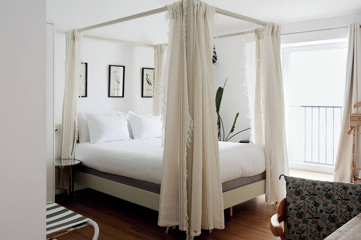 Una cama con dosel y cortinas, ropa de cama blanca, suelo de madera, una mesita de noche pequeña y vistas al balcón.