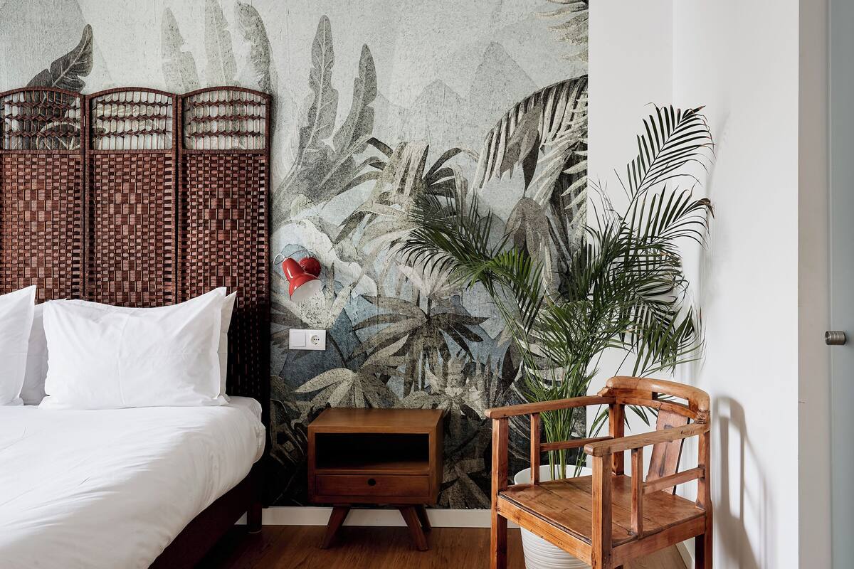 Un dormitorio con una cama, una mesita de noche, una silla de madera y una pared con un mural de hojas tropicales.