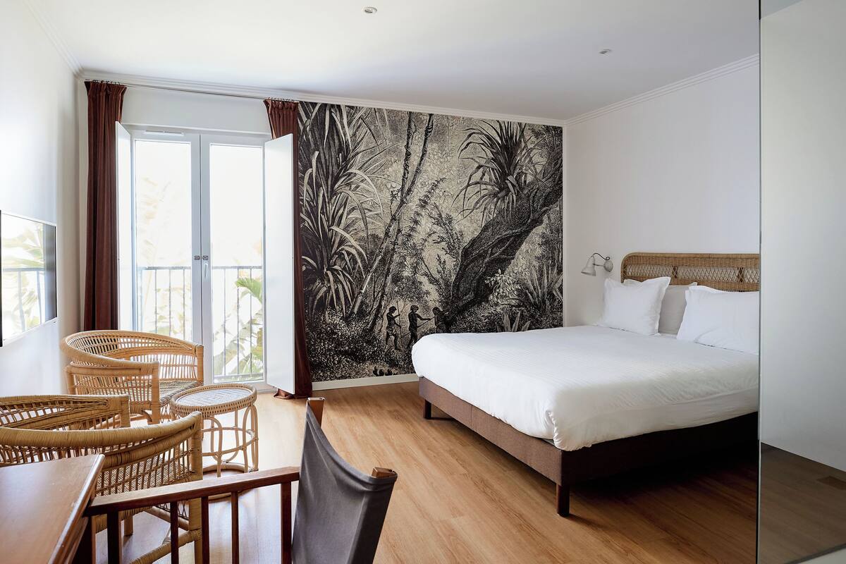 Un dormitorio con cama, una mesa de comedor con sillas, un ventanal grande y un mural en la pared.