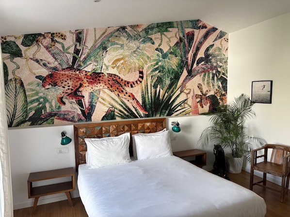 A bedroom with a large bed, a wooden headboard, a nightstand, a lamp, a chair, and a large botanical wall art piece. - Casa Apollo Guesthouse (Faro)
