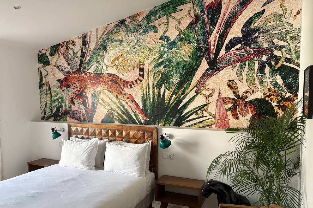 Un dormitorio con cama, mesitas de noche, una planta en maceta y un amplio mural tropical.