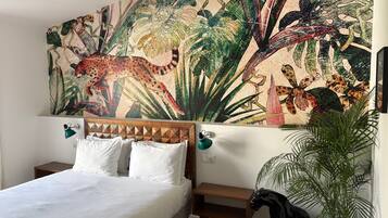 Un dormitorio con cama, mesitas de noche, una planta en maceta y un amplio mural tropical.