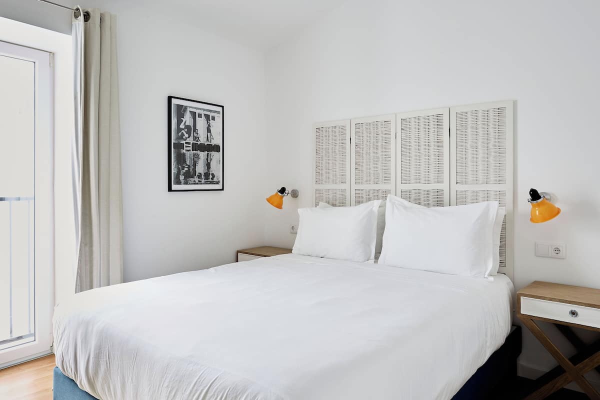 Una cama bien hecha con sábanas blancas, dos lámparas de noche, un cuadro enmarcado en la pared y una ventana con cortinas.