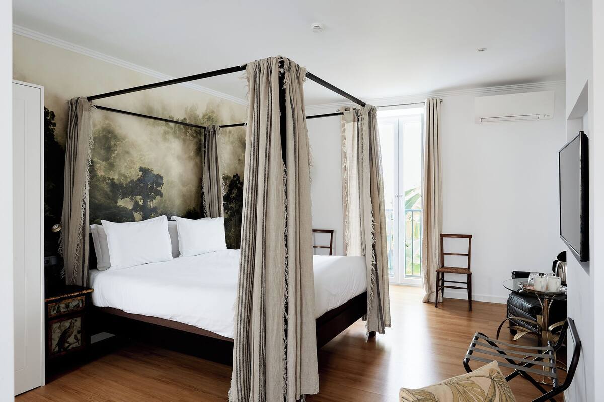 Una cama con dosel y cuatro columnas, con colchón y almohadas blancos, suelo de madera, una mesita con una bandeja de objetos y un televisor fijado a la pared.