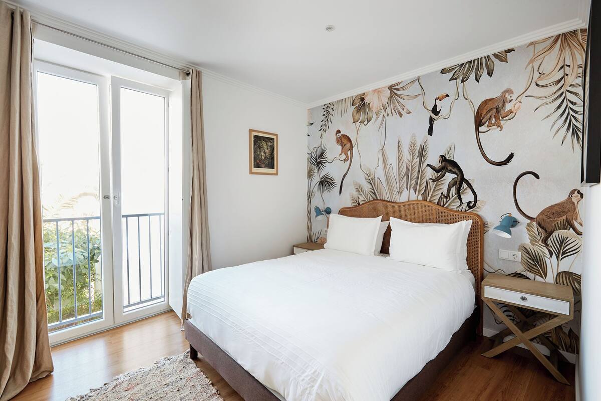 Un dormitorio con una cama grande, una mesita de noche de madera, una lámpara de pared y un ventanal con cortinas.