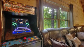 A gaming arcade with a racing game, a leather couch, and a window with a view of trees. - GOT IT ALL U ALL  Pool Table , Arcade ,Hot Tub , Pools,Free WIFI,Pet Friendly ! (Sevierville)