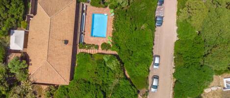 An aerial view of a house with a tiled roof, a swimming pool, and a driveway with cars parked.
