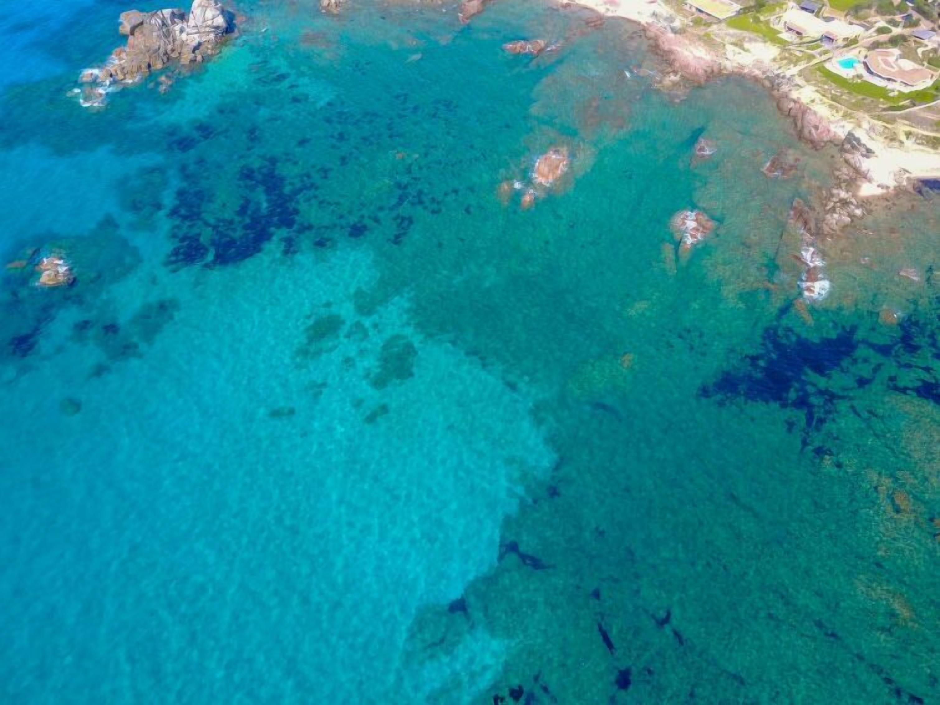 An aerial view of a coastal area with clear turquoise waters, rocky outcrops, and a developed beachfront with buildings and greenery.