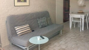 Um sofá cinzento com almofadas estampadas, uma pequena mesa redonda com um controle remoto e uma xícara, e uma mesa de jantar branca com cadeiras.